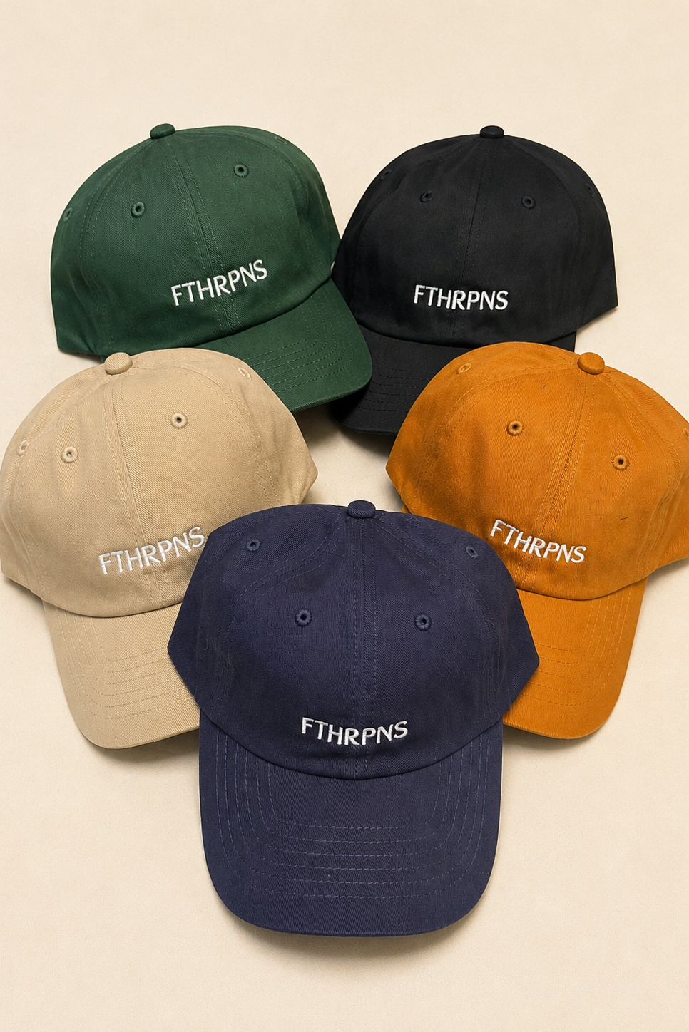 Five dad-style baseball caps arranged in a cluster on a light beige background. The caps are in green, black, beige, orange, and navy, each featuring the embroidered text 'FTHRPNS' on the front.