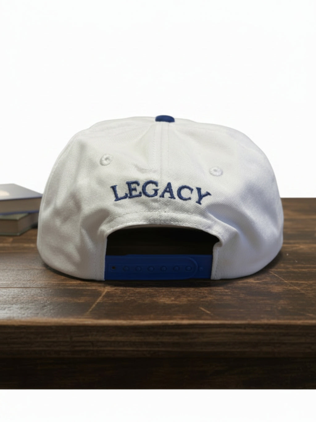 White baseball cap with blue embroidered 'LEGACY' text on the front, displayed on a dark wooden surface against a white background.
